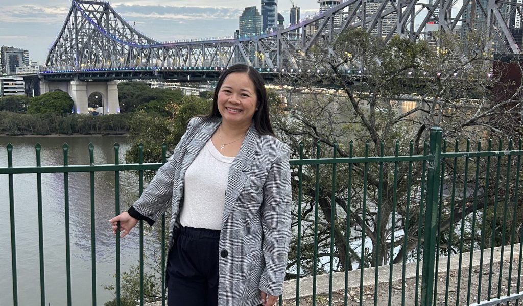 Southeast Asian woman standing in front an overlook of Brisbane bridge wearing black pants, white top, and patterened jacket. Smiling looking at the camera with hands by her side and holding onto the railing.